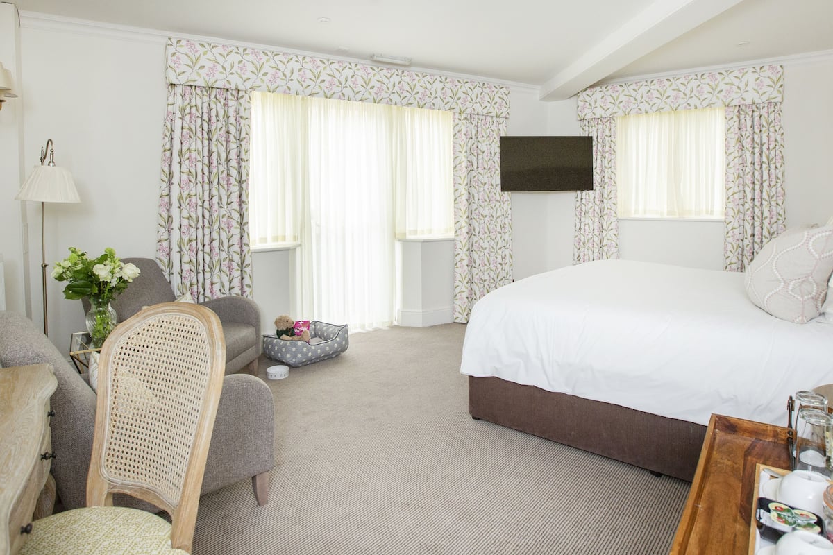 A hotel room with a large bed, a chair, a small table with items, and floral curtains.