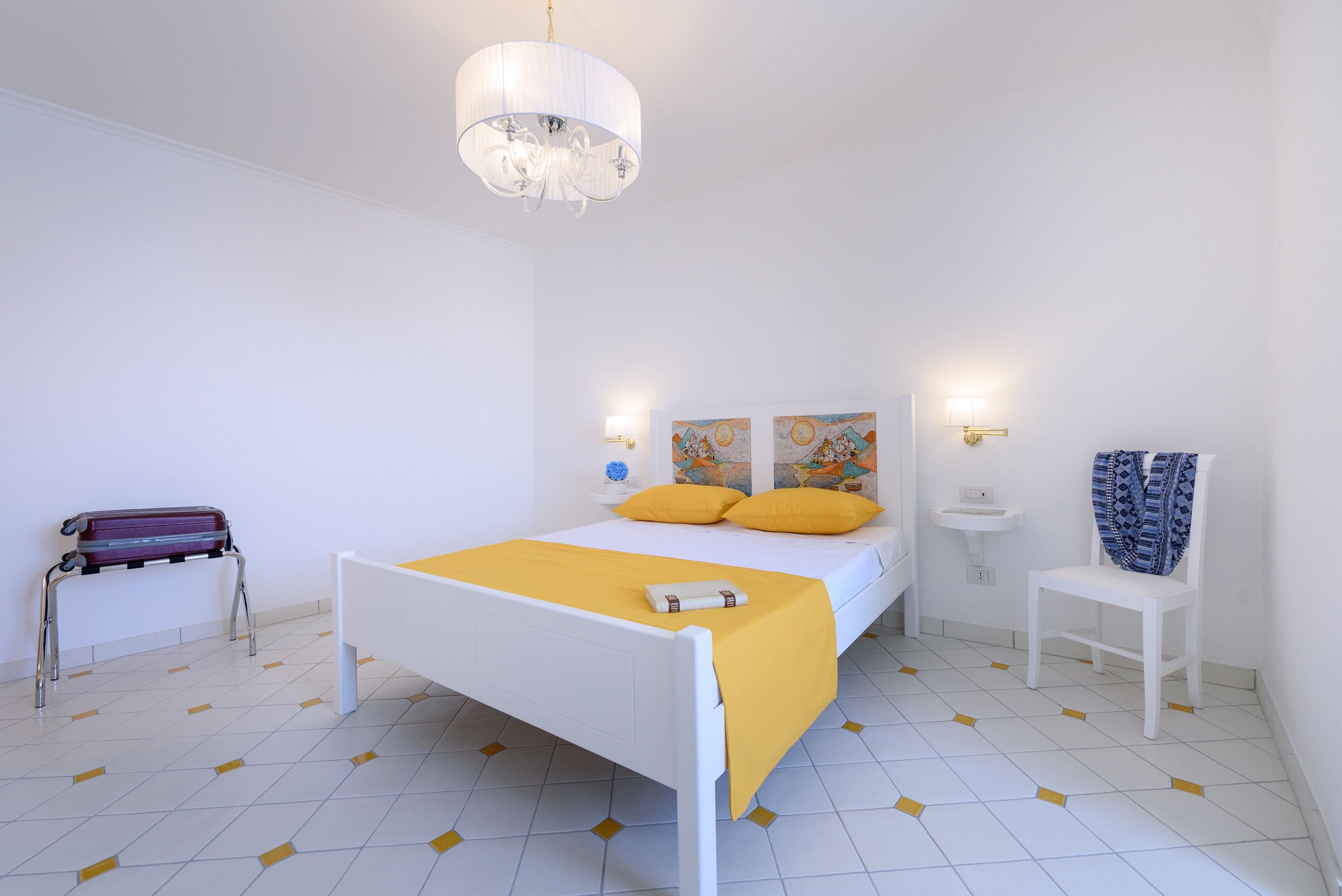 A bedroom with a bed, a bedside table, a chair, a suitcase, and a wall-mounted light fixture.