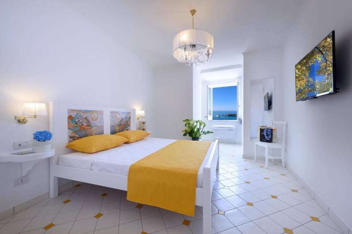 A modern hotel room with a white bed, yellow bedding, a chandelier, artwork on the walls, a TV, and a view of the sea through an open door.