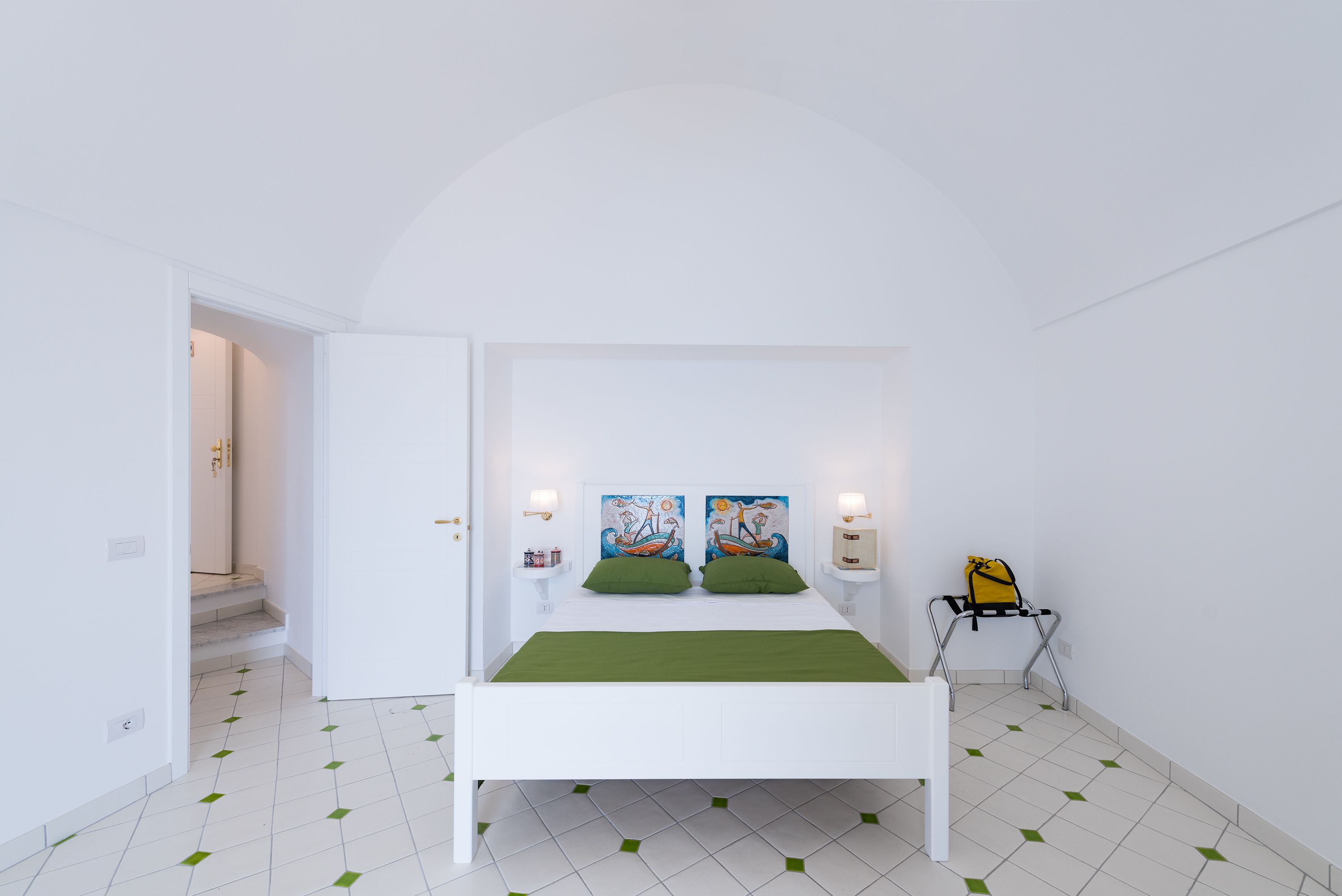 Une chambre à coucher blanche avec un lit, des oreillers verts, deux tableaux accrochés au mur, une table de chevet avec une lampe et une valise posée sur une chaise.
