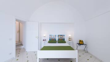 Une chambre à coucher blanche avec un lit, des oreillers verts, deux tableaux accrochés au mur, une table de chevet avec une lampe et une valise posée sur une chaise.