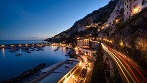Un pueblo costero al anochecer con un puerto deportivo, edificios a lo largo del acantilado y una carretera con estelas de luz.