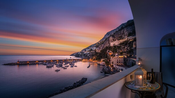 Un balcón con vistas a un pueblo costero al atardecer, mostrando un mar tranquilo, barcos y edificios a lo largo del acantilado.