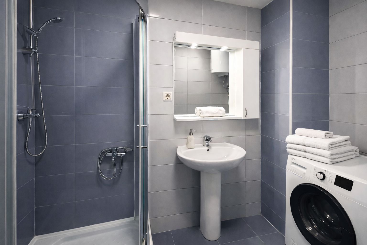 A modern bathroom with a glass shower enclosure, a pedestal sink, a mirror above the sink, and a stacked washer and dryer.