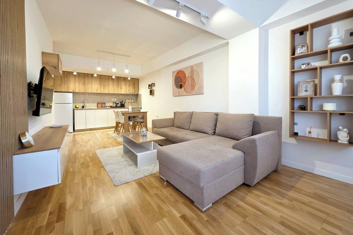 A modern living room with a large sectional sofa, a kitchen in the background, and a wooden floor.