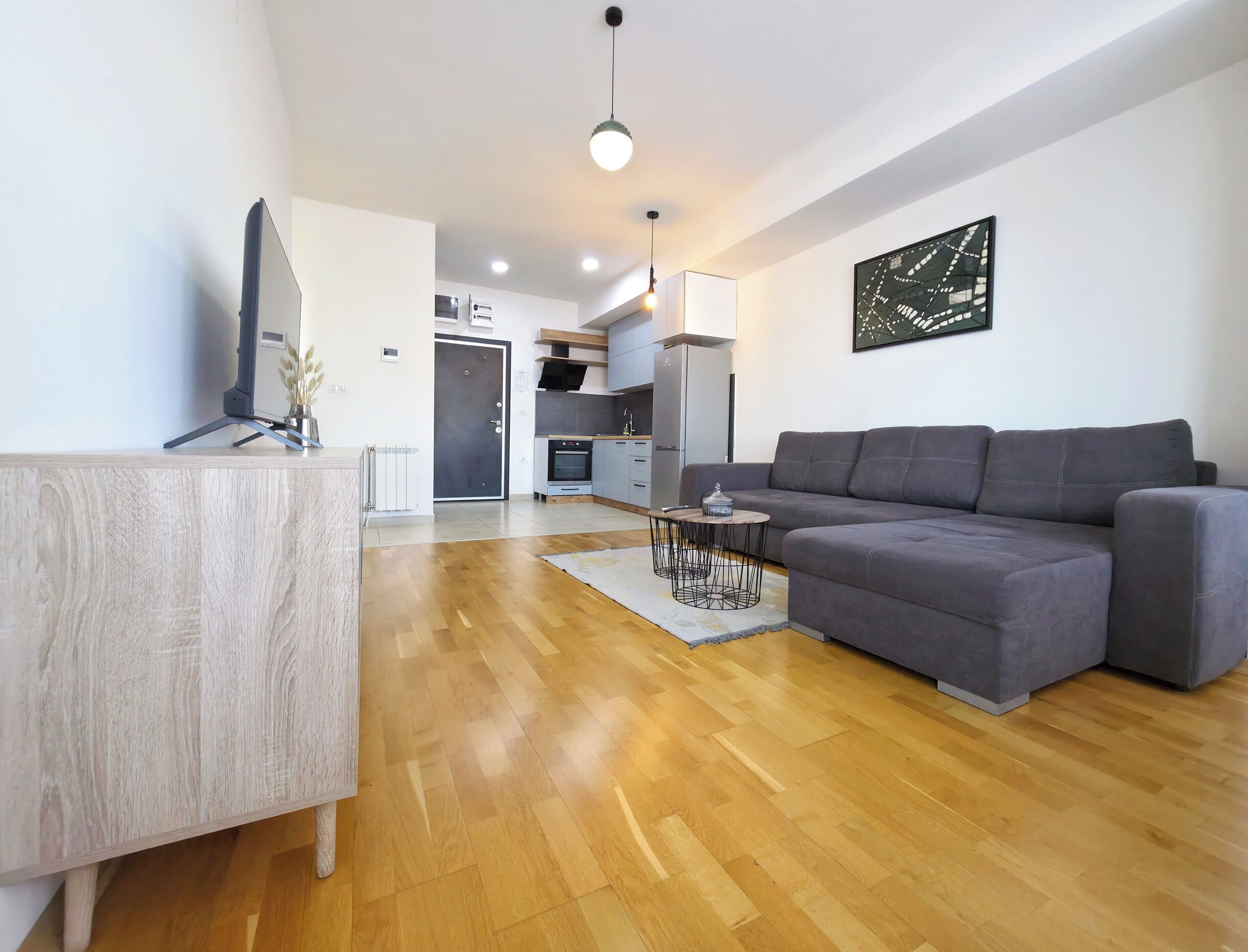 Ein modernes Wohnzimmer mit einer grauen Couch, Holzboden, einem kleinen runden Tisch und einer Küche im Hintergrund.