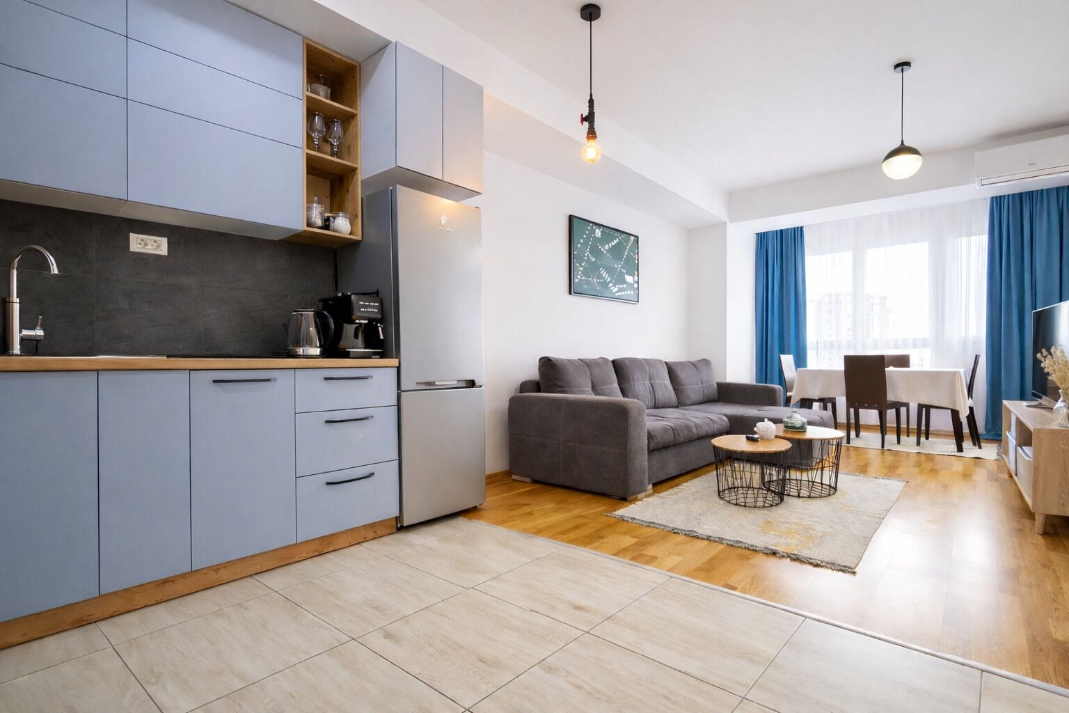 A modern kitchen with grey cabinets, a built-in oven, and a coffee machine. There is a grey sofa, a dining table with chairs, and a TV in the living area.