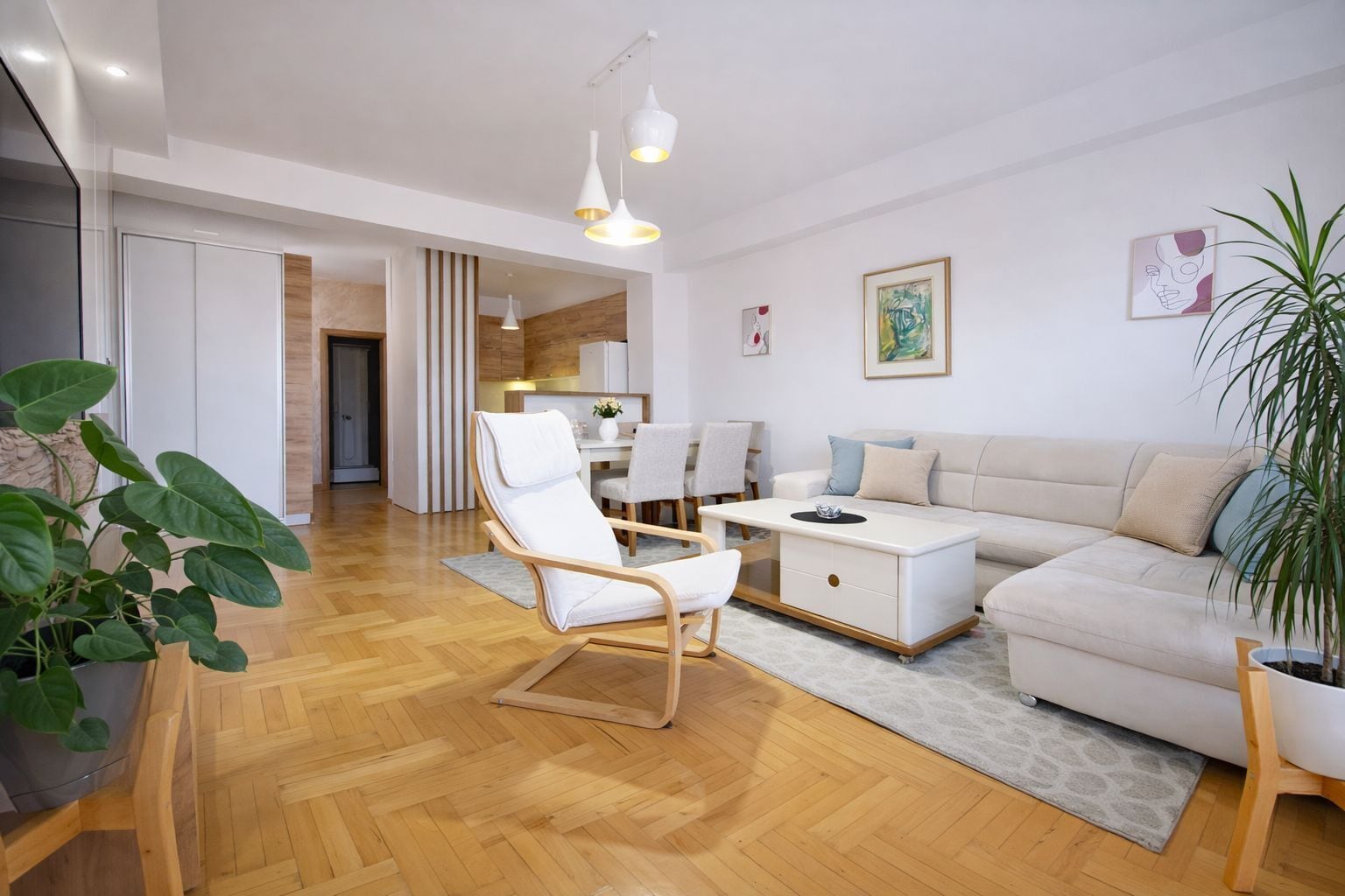A modern living room with a wooden floor, a white sofa, a white armchair, and a dining area in the background.