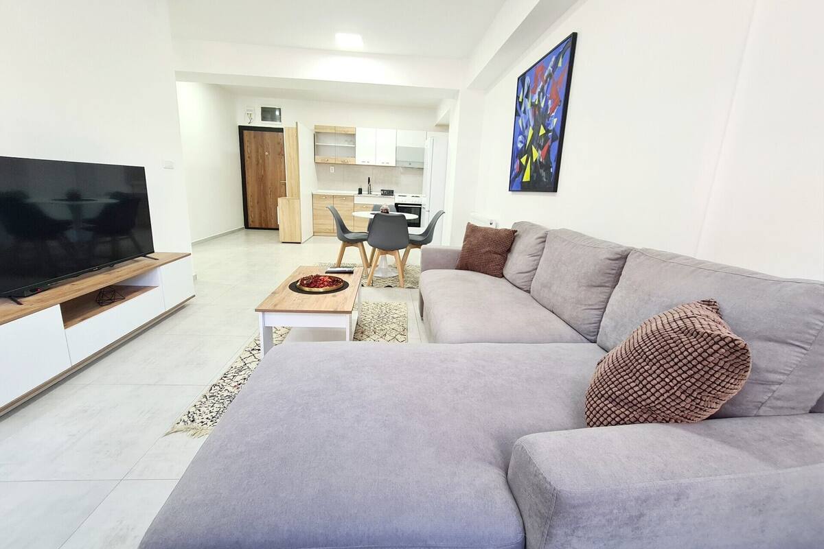 A modern living room with a grey sofa, a flat-screen TV, a dining area with a table and chairs, and a kitchen in the background.