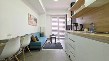 Ein kompakter Wohnraum mit Küchenzeile, einer Couch, einem Essbereich und Blick durch ein Fenster mit Jalousien.