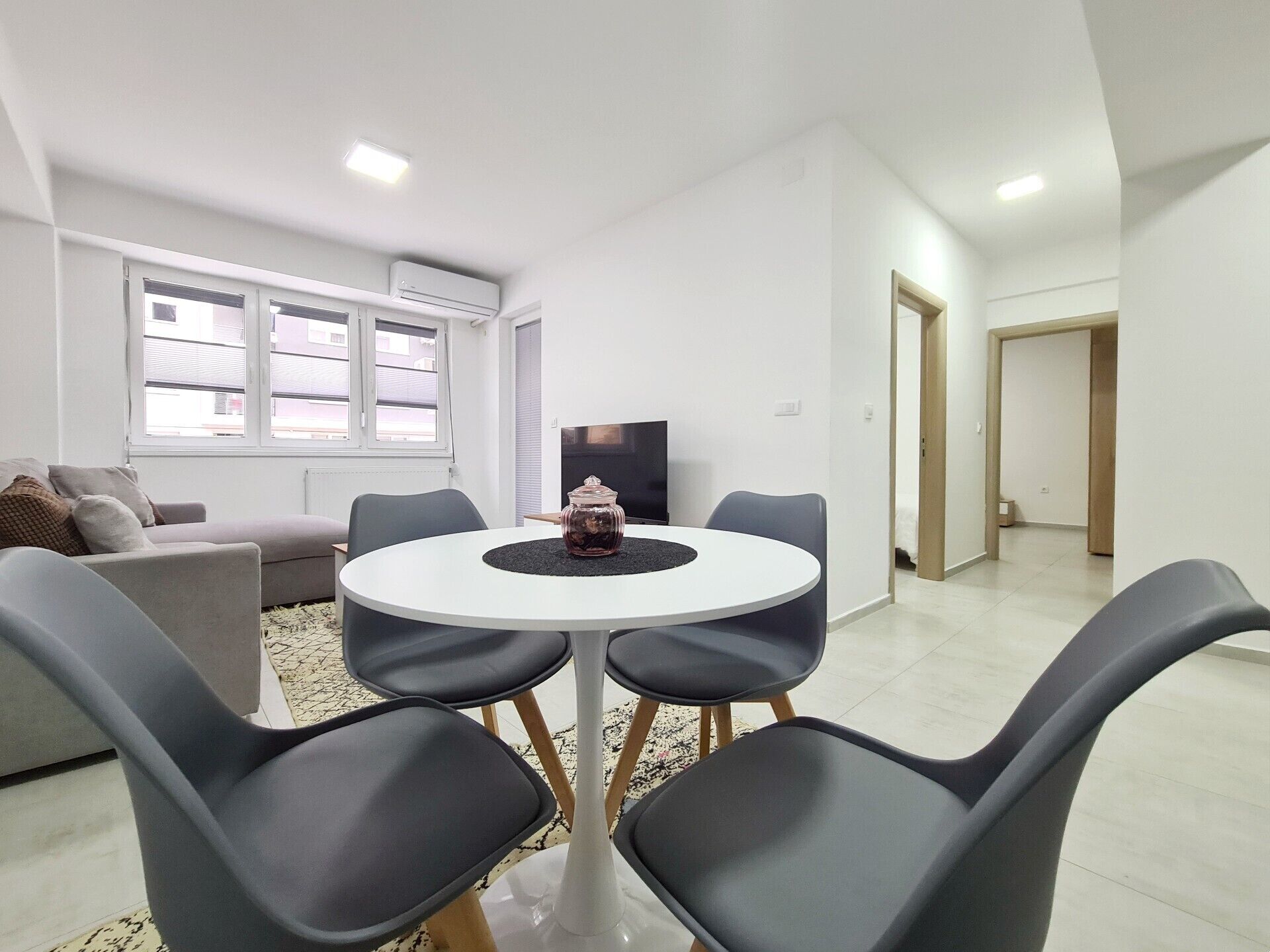 A modern living room with a round table, grey chairs, a sofa, a TV, and a window with blinds.