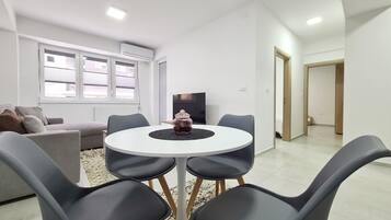 A modern living room with a round table, grey chairs, a sofa, a TV, and a window with blinds.
