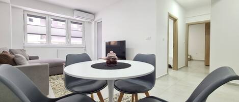A modern living room with a round table, grey chairs, a sofa, a TV, and a window with blinds.