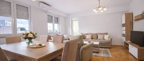 A modern living room with a wooden dining table, beige sofa, and a TV on a wooden stand.