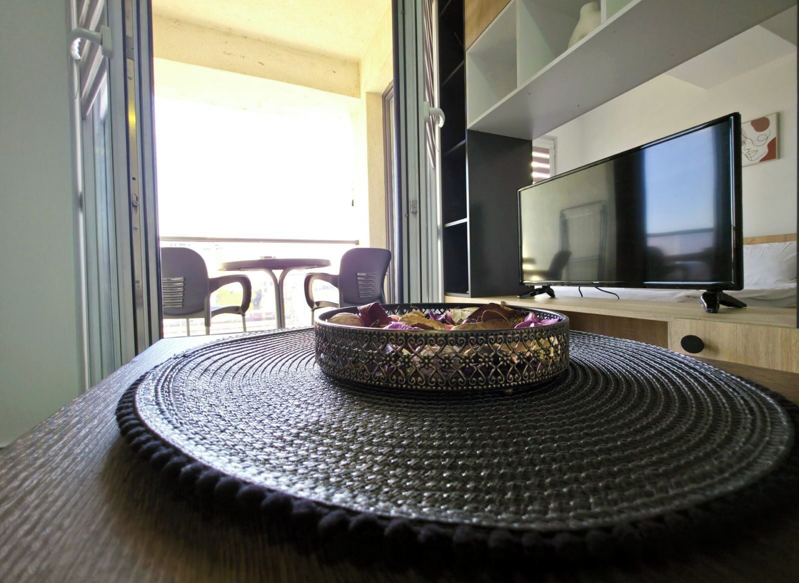 Une pièce avec un téléviseur posé sur un meuble en bois, un plateau décoré sur un tapis tissé, et une vue sur le balcon extérieur avec ses chaises.
