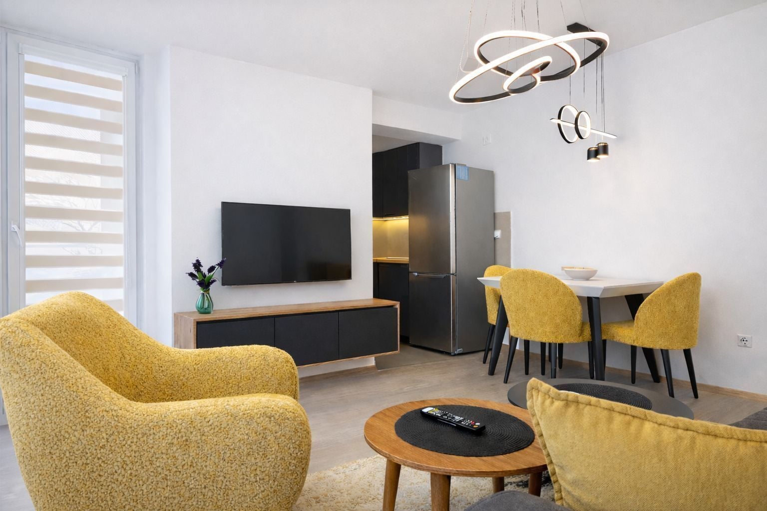 Uma sala de estar moderna com televisão de tela plana, cadeiras amarelas, uma mesa de jantar e uma área de cozinha com geladeira.