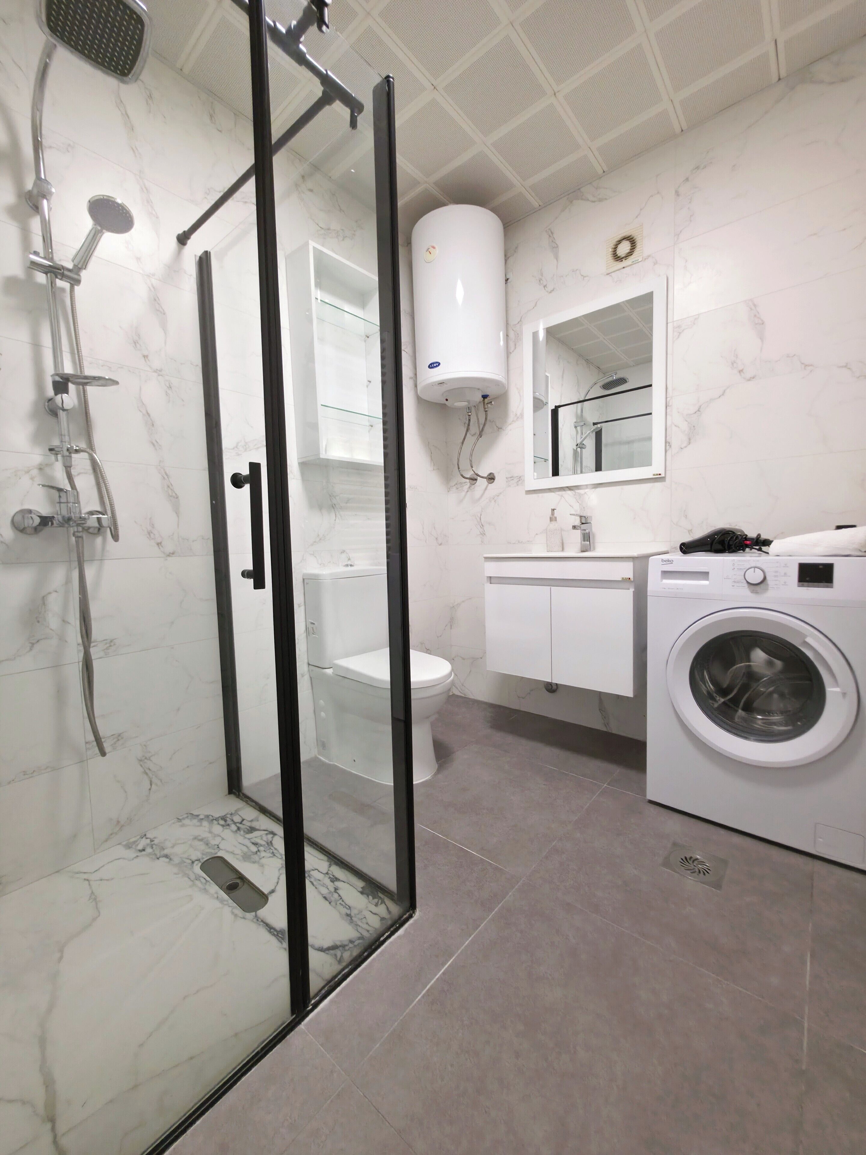 A bathroom with a glass-enclosed shower, a toilet, a sink with a mirror, and a washing machine.