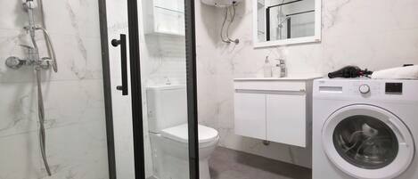 A bathroom with a glass-enclosed shower, a toilet, a sink with a mirror, and a washing machine.