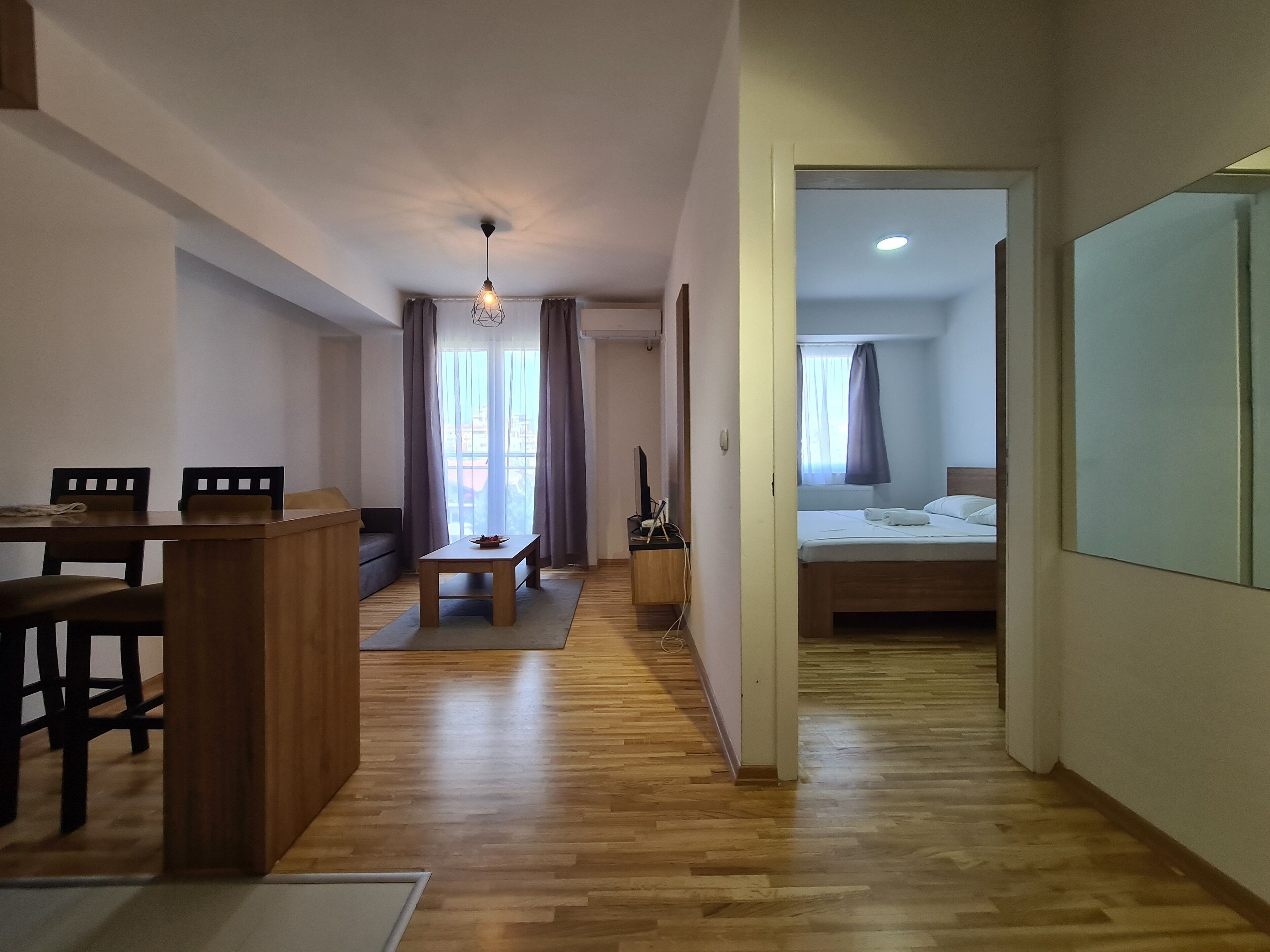 A modern hotel room with a wooden floor, a dining area with a table and chairs, a sofa, a bed with a white mattress, and a large mirror on the wall.