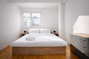 Une chambre à coucher moderne avec un cadre de lit en bois, une literie blanche et une table de chevet en bois surmontée d’une lampe.