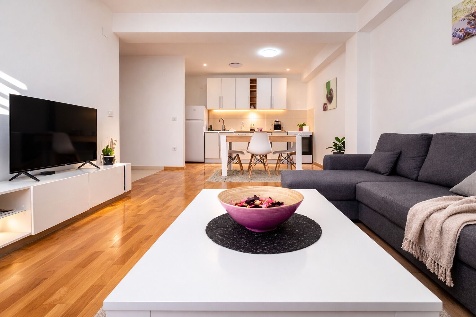 A modern living room with a flat-screen TV, a grey sofa, and a white coffee table with a purple bowl.