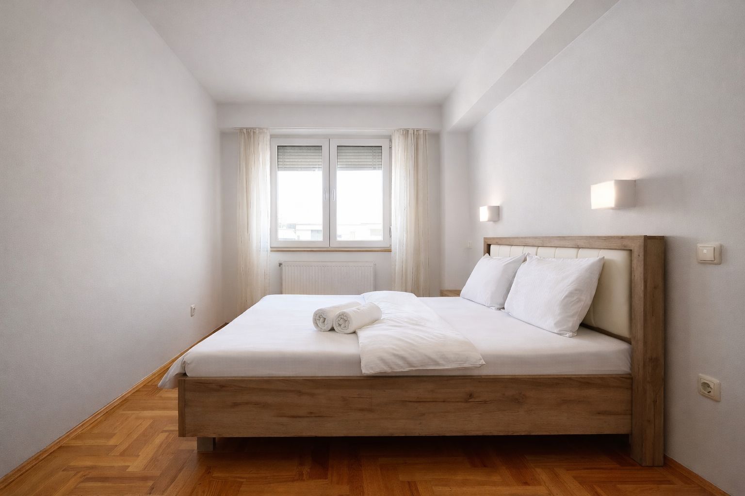 A bedroom with a wooden bed, white bedding, a window with curtains, and a radiator.