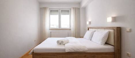 Un dormitorio con una cama de madera, ropa de cama blanca, una ventana con cortinas y un radiador.
