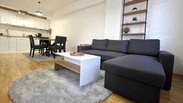 Una sala de estar moderna con un sofá gris, una mesa de centro blanca y un comedor con sillas, con una cocina al fondo.
