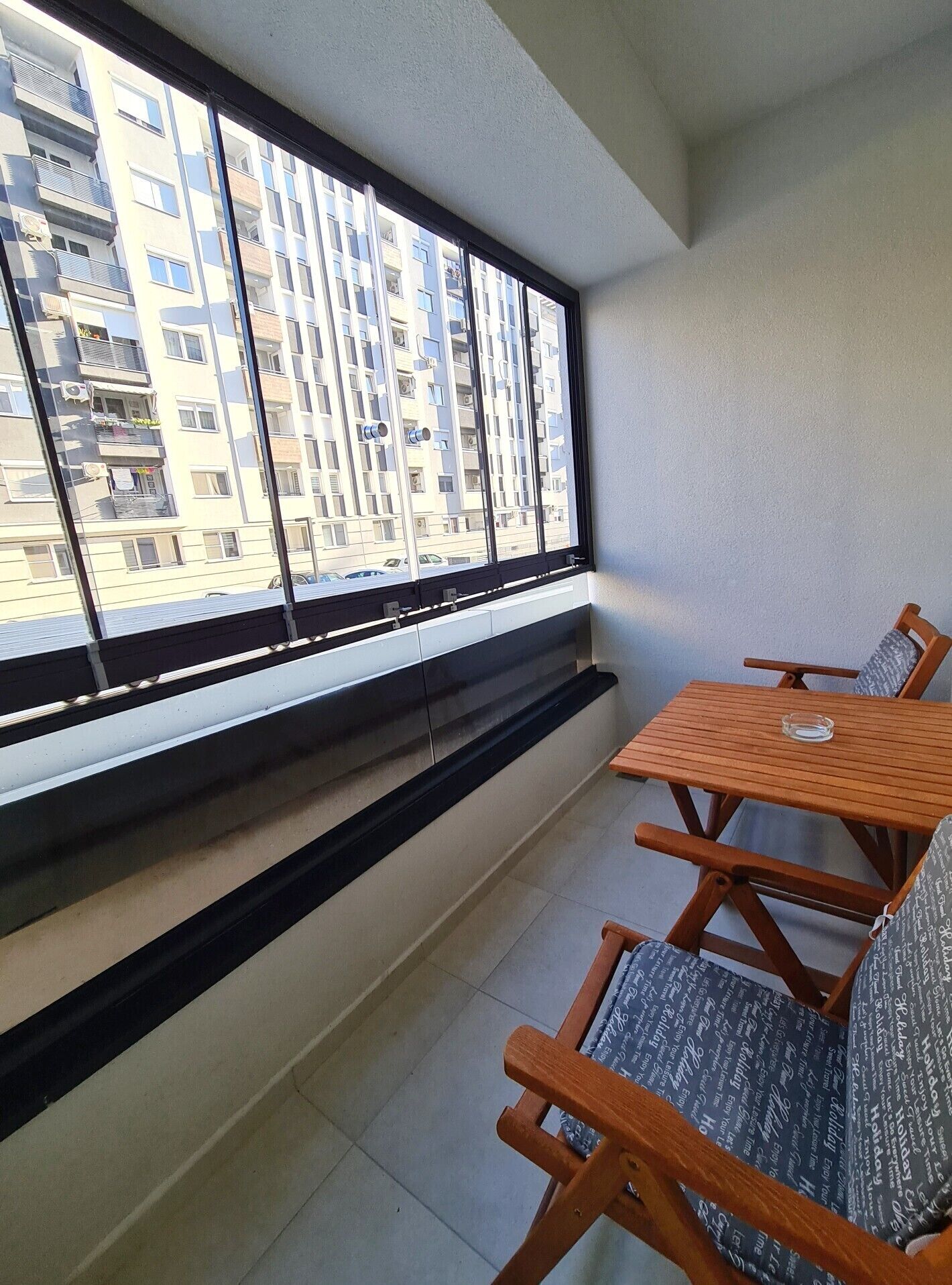 A balcony with a view of a residential building, a wooden table, and two chairs.