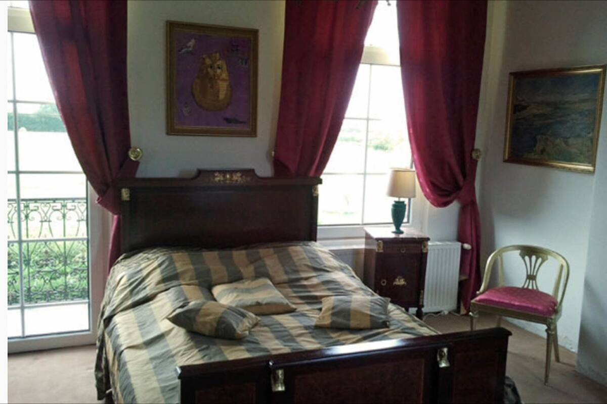 Een slaapkamer met een houten bed, rode gordijnen, een raam met uitzicht, een radiator en een stoel.