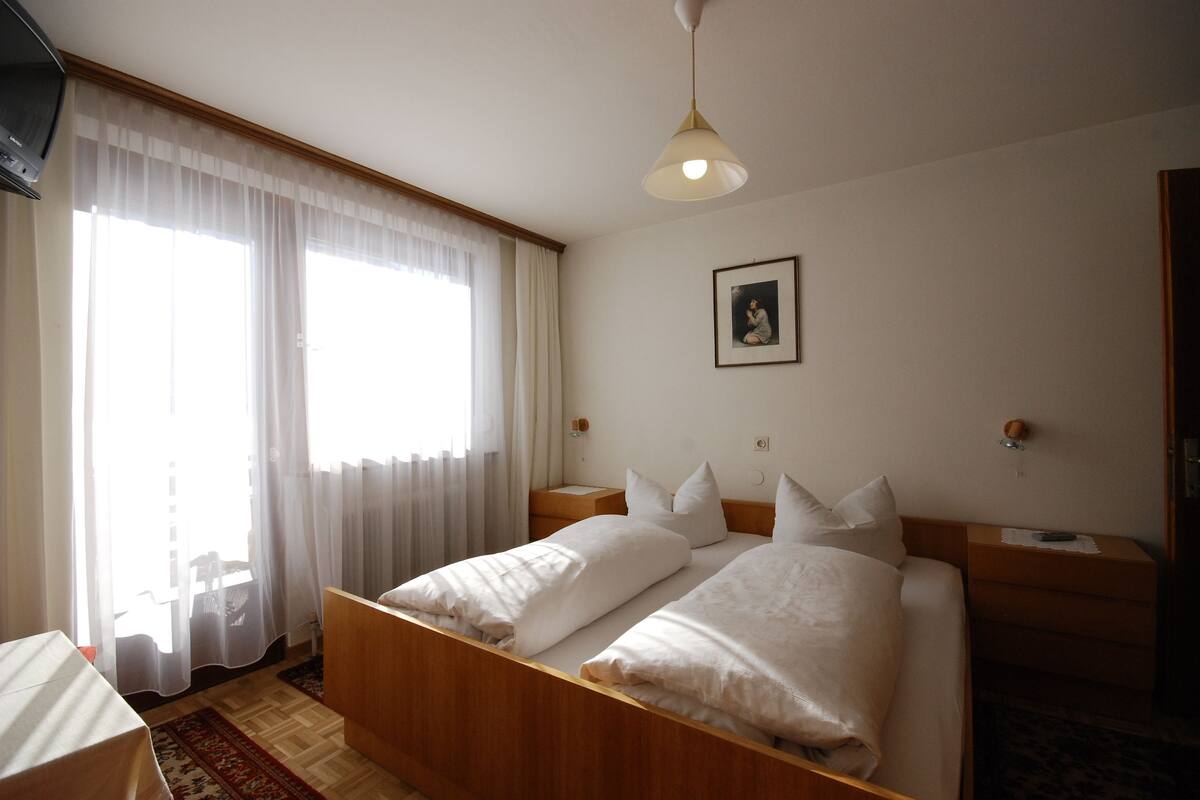 A bedroom with a wooden bed, white bedding, a bedside table, a nightstand, a framed picture on the wall, and a window with sheer curtains.