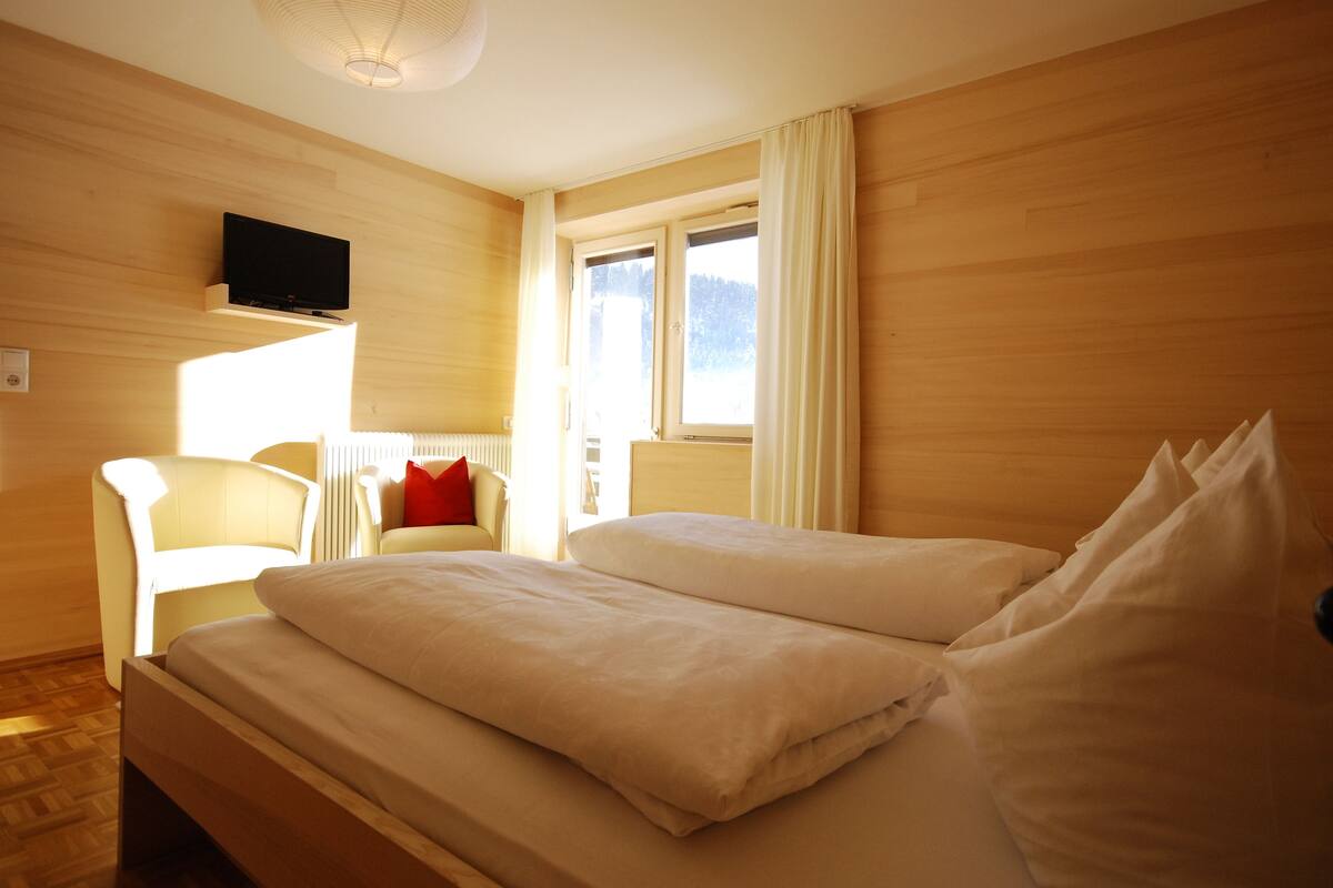 A hotel room with a wooden floor, a bed with white bedding, a television mounted on the wall, a window with curtains, and a chair with a red pillow.