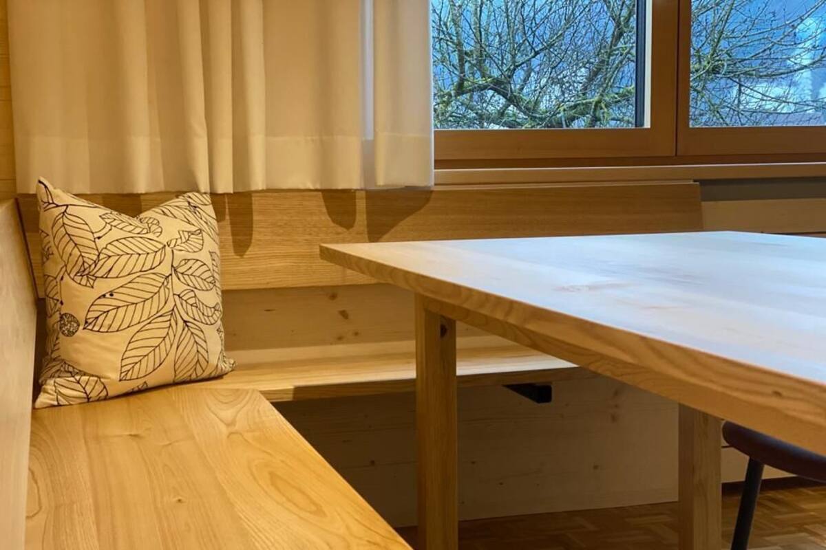 A wooden bench with a leaf-patterned pillow, a wooden table, and a window with a view of trees.