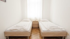 Una habitación pequeña y limpia con dos camas individuales, una mesita de noche y una ventana con cortinas traslúcidas.