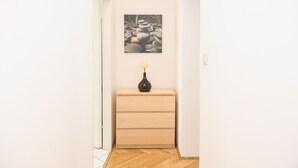 Un pasillo con piso de madera, un aparador beige con cajones, un florero con una flor y un cuadro de rocas en la pared.