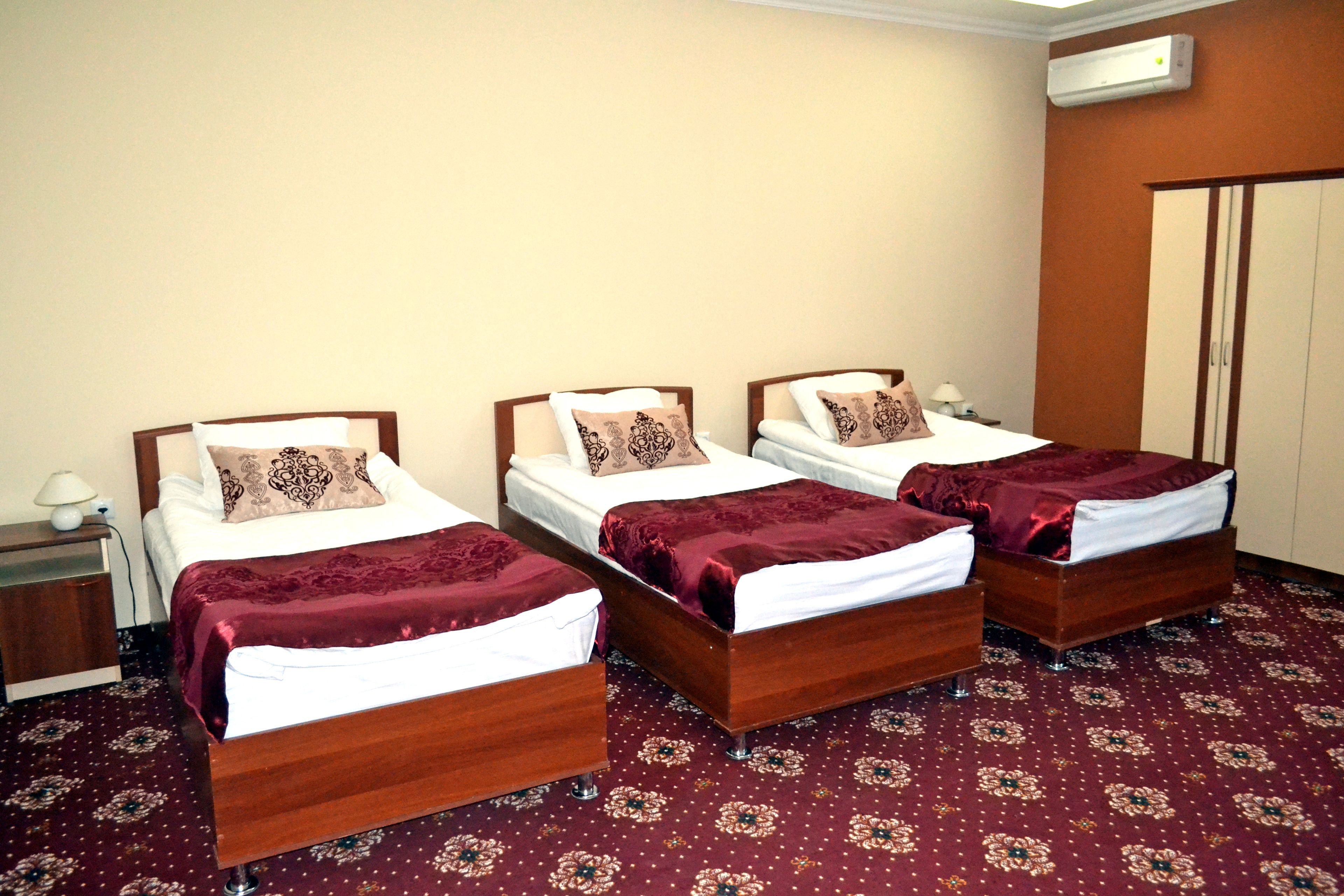 A hotel room with three wooden beds, each with a white mattress and a brown headboard. The room has a patterned carpet and an air conditioning unit on the wall.