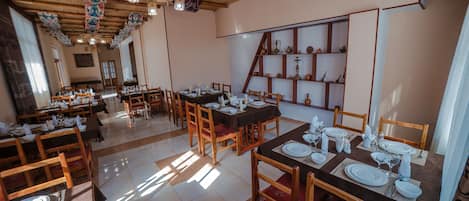 Un restaurante con varias mesas dispuestas para recibir comensales, sillas de madera y piso de baldosas.