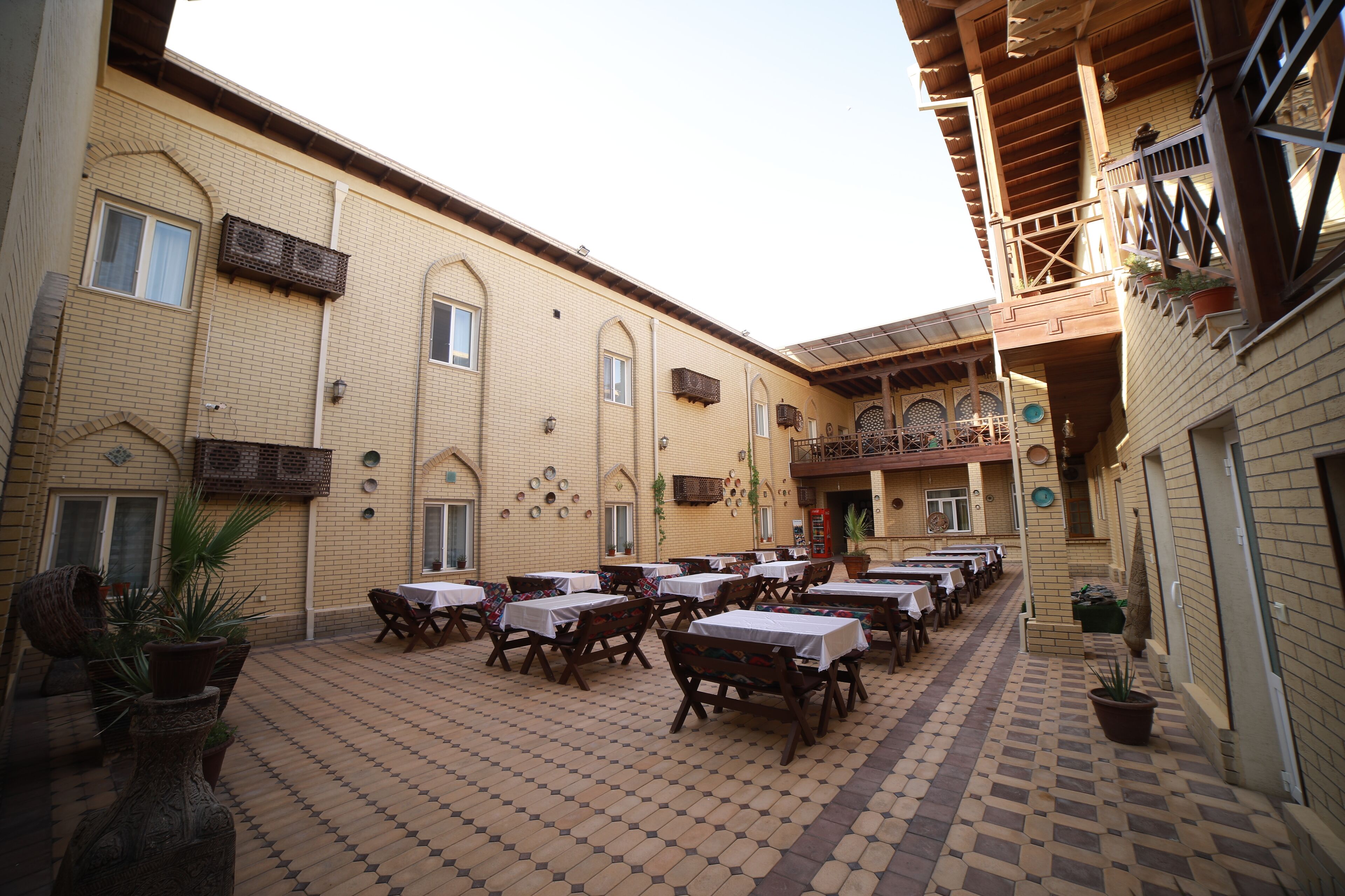 Un espacio de comedor al aire libre con varias mesas y sillas, un edificio de ladrillo con ventanas de arco y un piso de baldosas.