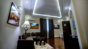 A living room with a sofa, a table, and a lamp. There is a painting on the wall and a window with curtains. - Arkanchi Hotel (Khiva)