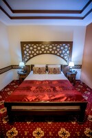 Una cama bien hecha con una colcha roja, dos lámparas de noche y un cabecero decorativo.