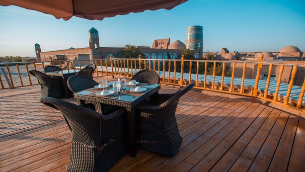 A wooden deck with wicker furniture and a table set for dining, overlooking a historic building with domes and towers. - Arkanchi Hotel (Khiva)