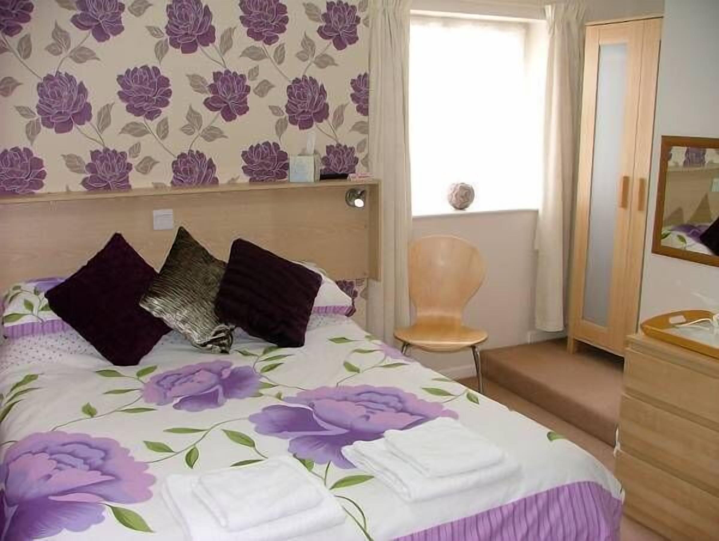 Une chambre à coucher avec une couvre-lit à motifs floraux, une tête de lit en bois, une chaise, une armoire et une fenêtre avec des rideaux.