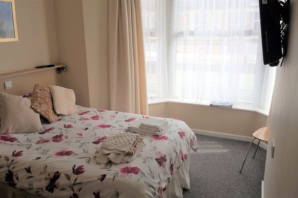 A hotel room with a bed, pillows, a window with curtains, and a small table.