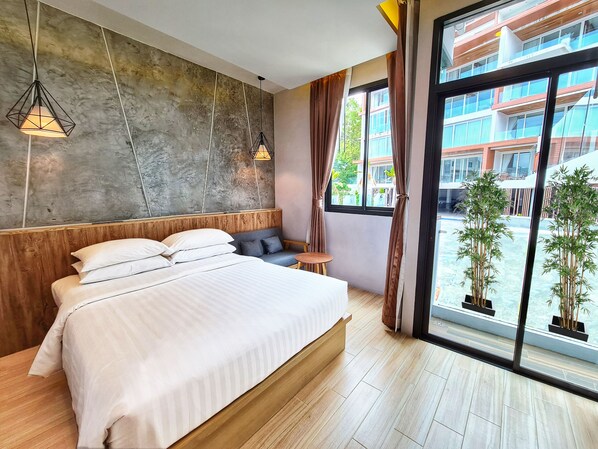 A modern bedroom with a large bed, wooden headboard, and a view of a building through large windows. - Oneloft Hotel (Karon)
