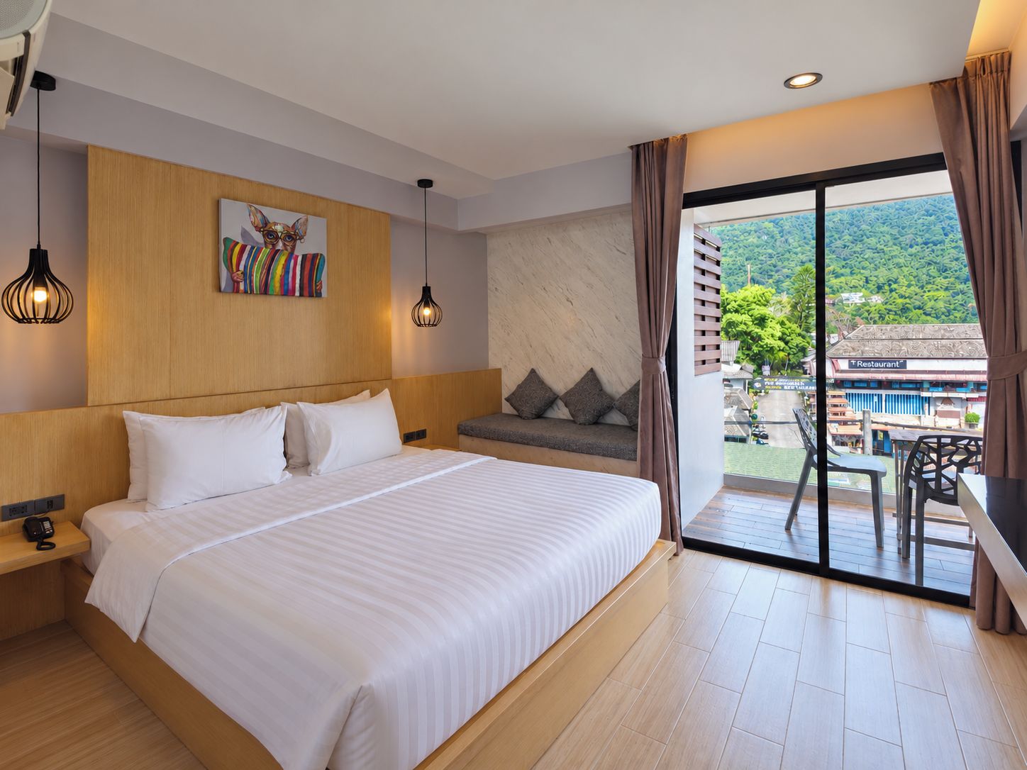 A hotel room with a large bed, a sofa, a painting, and a view of a restaurant and greenery outside.