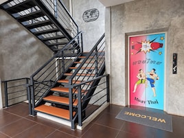 Una escalera con barandillas negras y peldaños de madera, que lleva a una puerta con un cartel llamativo de luchadores de Muay Thai.