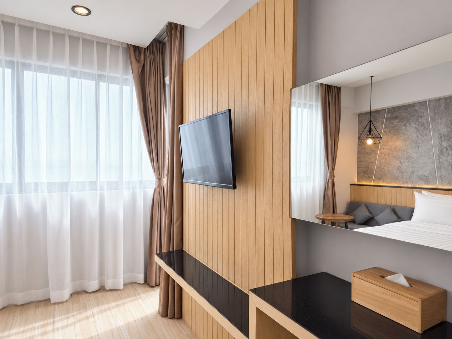 Habitación de hotel moderna con televisión de pantalla plana montada en la pared, un panel decorativo de madera, un espejo grande, una cama con almohadas y una mesita de noche con un dispensador de pañuelos.