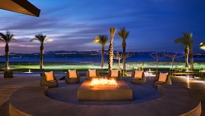 A circular outdoor seating area with a fire feature, surrounded by lounge chairs and palm trees, overlooking a body of water and distant city lights. - The Setai Sea of Galilee (Ramot)