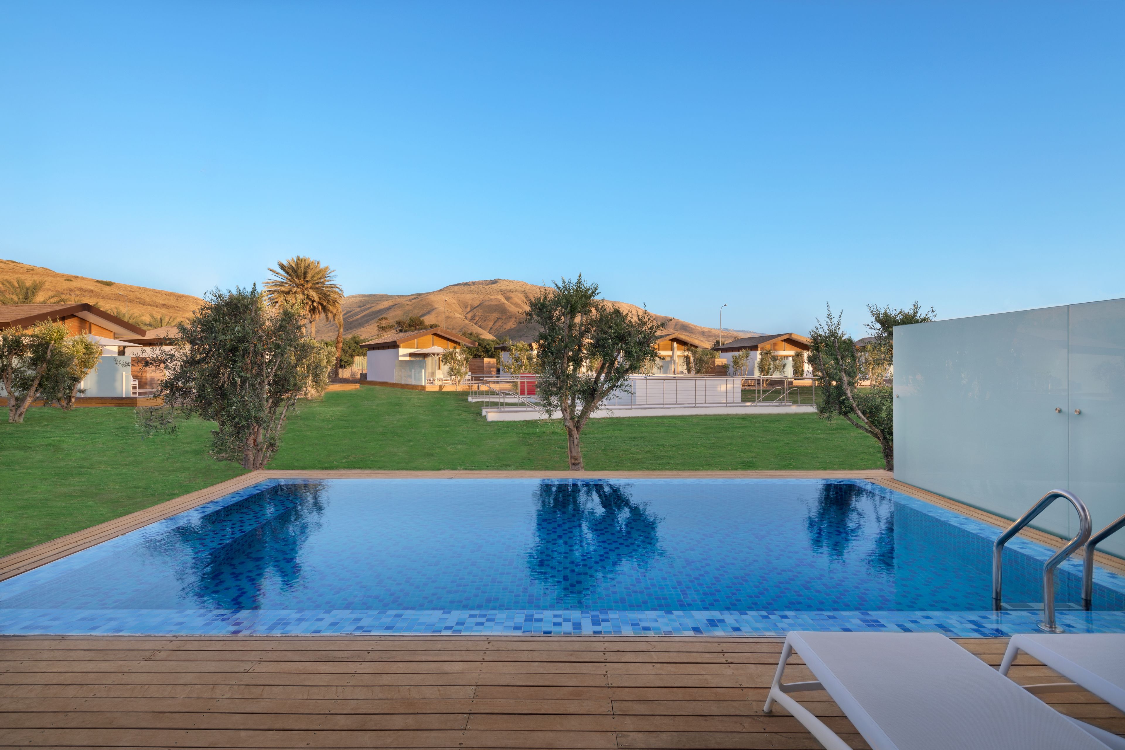 Una piscina con cubierta de madera, rodeada de césped y palmeras, con casas al fondo.