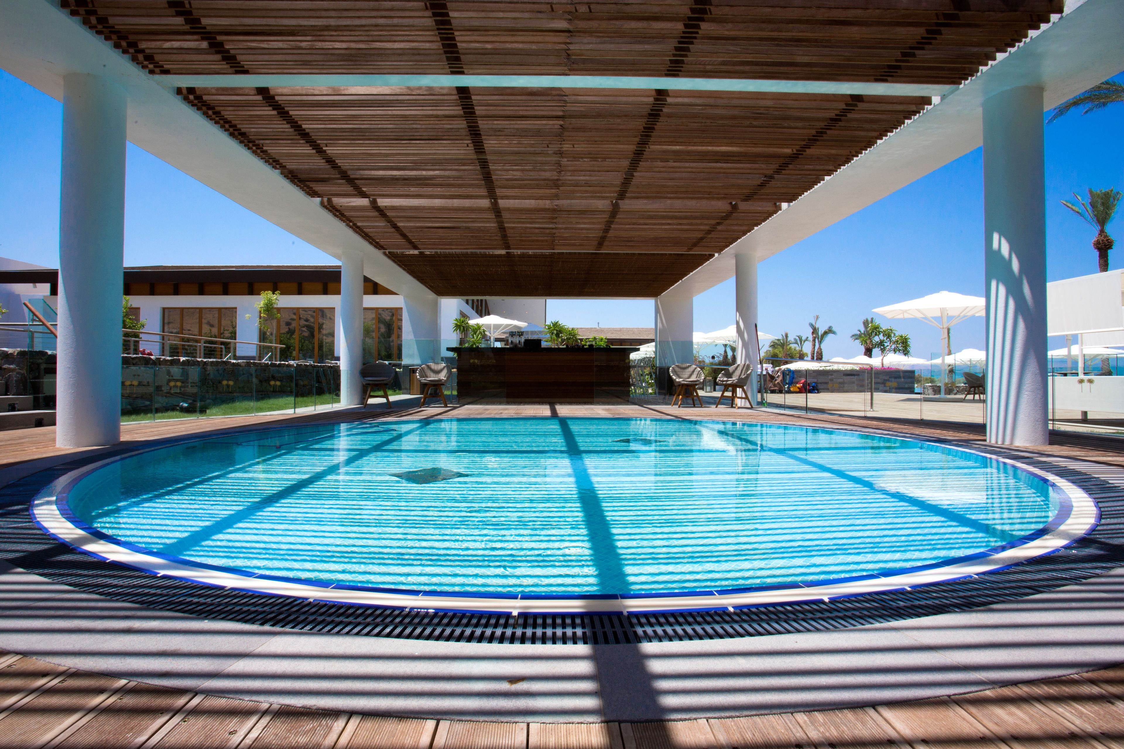 Una piscina con cubierta de madera, rodeada de columnas blancas y sombrillas, bajo una pérgola de madera.